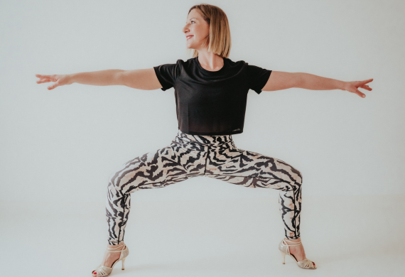 Frau in einer Yoga-Pose, trägt ein schwarzes Oberteil und gemusterte Leggings und steht in High Heels auf einem weißen Hintergrund.